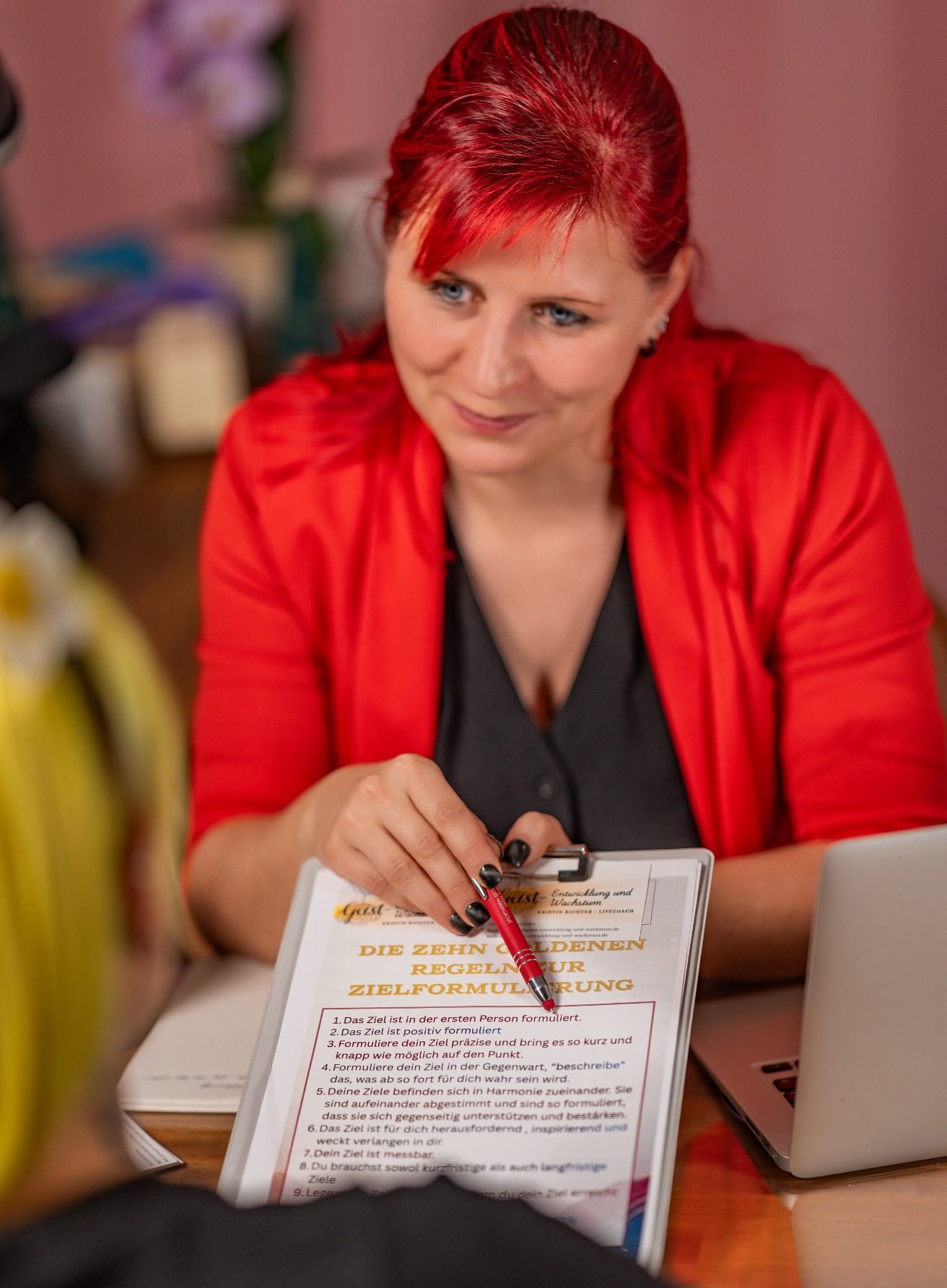 Eine Person mit rotem Blazer hält ein Notizbuch und einen Stift in der Hand.